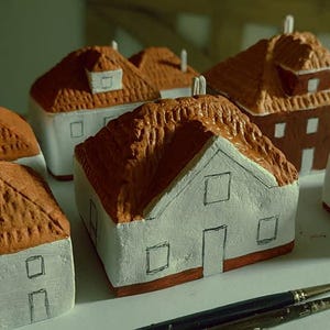 Może przedstawiać: Kolekcja sześciu miniaturowych domk&oacute;w wykonanych z gliny, pomalowanych na biało z brązowymi dachami. Domki mają małe okna i drzwi, a niekt&oacute;re mają kominy. Domki są ułożone na białej powierzchni.
