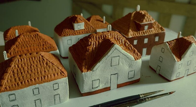 Może przedstawiać: Kolekcja sześciu miniaturowych domk&oacute;w wykonanych z gliny, pomalowanych na biało z brązowymi dachami. Domki mają małe okna i drzwi, a niekt&oacute;re mają kominy. Domki są ułożone na białej powierzchni.