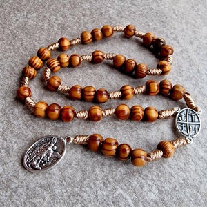 May include: A wooden rosary with brown beads and a silver pendant featuring a religious image. The rosary is strung with a beige cord.