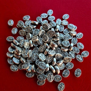 Puede incluir: Un montón de cuentas ovaladas plateadas con un diseño en relieve. Las cuentas están esparcidas sobre una superficie roja.