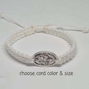 May include: White braided cord bracelet with a silver oval charm featuring a depiction of St. Francis. The charm has the text "ST. FRANCIS" inscribed on it.  Choose cord color and size.