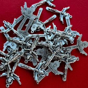 May include: A collection of silver-toned metal charms featuring a cross design. Each charm has a different inscription in a stylised font. The charms are arranged in a pile on a red surface.