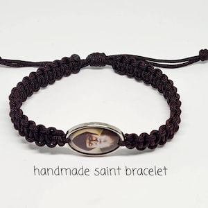 May include: A brown braided bracelet with a silver oval charm featuring a picture of a saint. The bracelet is handmade and has the text "handmade saint bracelet" below the image.