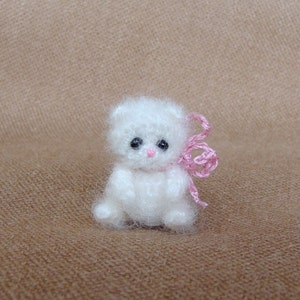 May include: A small white crocheted teddy bear with black eyes and a pink bow.