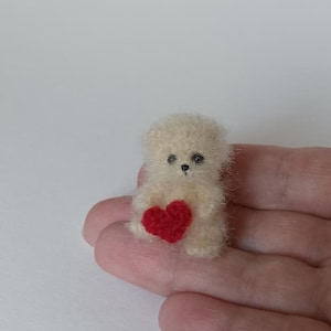 Puede incluir: Un pequeño oso de peluche blanco tejido a crochet sosteniendo un corazón rojo. El oso está sentado en una mano.