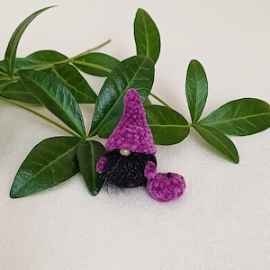 May include: A small purple crocheted gnome with a black fuzzy beard and a purple crocheted pumpkin. The gnome is sitting on a white surface with green leaves behind it.