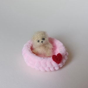 Puede incluir: Un pequeño perro marrón claro y esponjoso se sienta en una cama rosa en forma de corazón con un corazón rojo cosido en el lateral.
