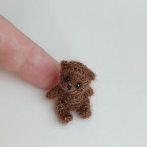 May include: A small brown crocheted teddy bear with black eyes. The bear is sitting on a white surface.