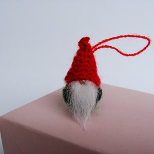 May include: A small red knitted gnome ornament with a white beard and a green felt base. The gnome has a red string loop for hanging.