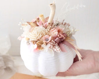 May include: A white pumpkin with a stem made of natural wood is filled with dried flowers in shades of pink, cream, and brown. The pumpkin is sitting on a brown envelope with a heart-shaped detail.
