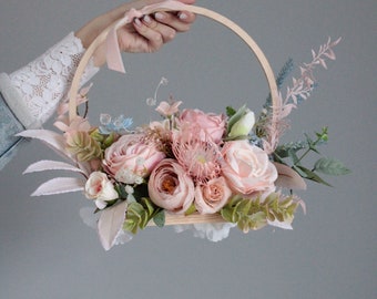 Op de afbeelding: Een bloemstuk met een houten hoepel versierd met roze en witte bloemen, groen blad en blauwe accenten. Bovenop is een roze lint vastgebonden. De opstelling wordt door een persoon vastgehouden.