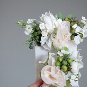 May include: A white canvas with a floral arrangement of white and cream flowers and green leaves. The flowers are arranged in a cluster and are attached to the canvas.