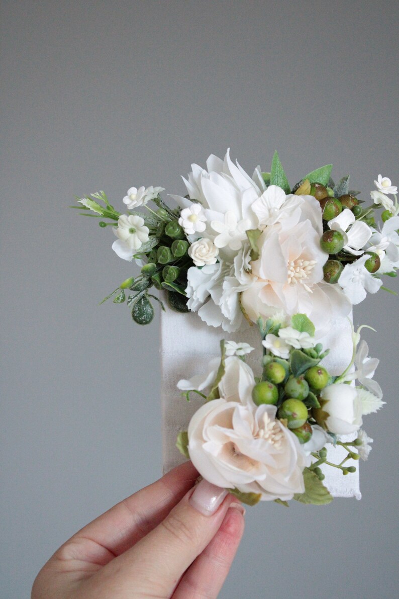 May include: A white canvas with a floral arrangement of white and cream flowers and green leaves. The flowers are arranged in a cluster and are attached to the canvas.