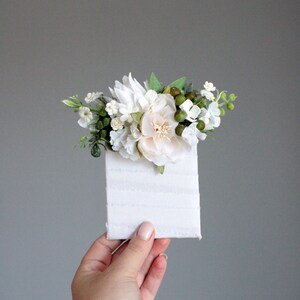 May include: A small white card with a white and green floral arrangement on top. The flowers are mostly white with a few light pink flowers.