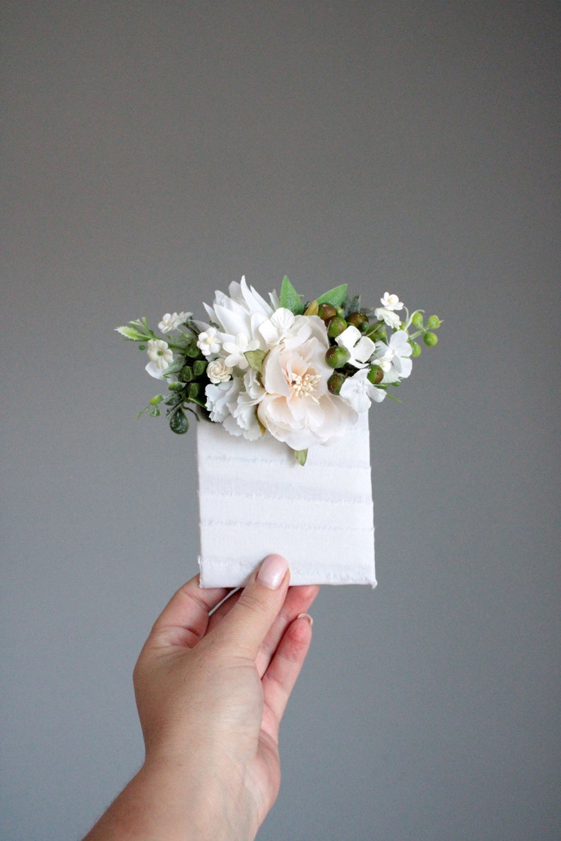 May include: A small white card with a white and green floral arrangement on top. The flowers are mostly white with a few light pink flowers.