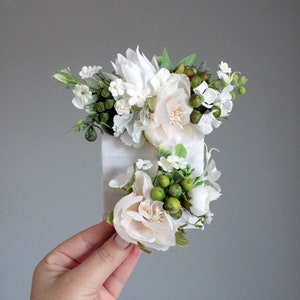 May include: A white and cream floral hair comb with green berries and leaves. The comb is made of metal and has a white ribbon attached to it.