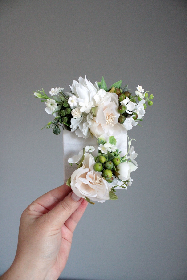 May include: A white and cream floral hair comb with green berries and leaves. The comb is made of metal and has a white ribbon attached to it.