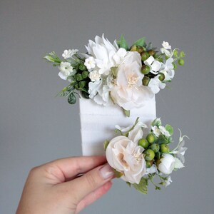 May include: Two white flower hair clips with green leaves and berries. The clips are on a white background.