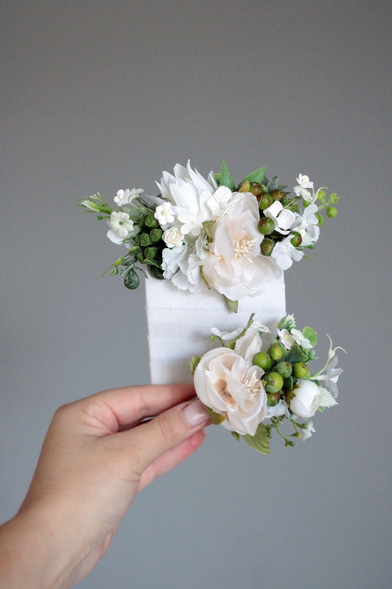 May include: Two white flower hair clips with green leaves and berries. The clips are on a white background.