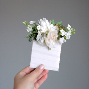 May include: A small bouquet of white flowers with green leaves and berries, arranged on a white card with a textured surface.
