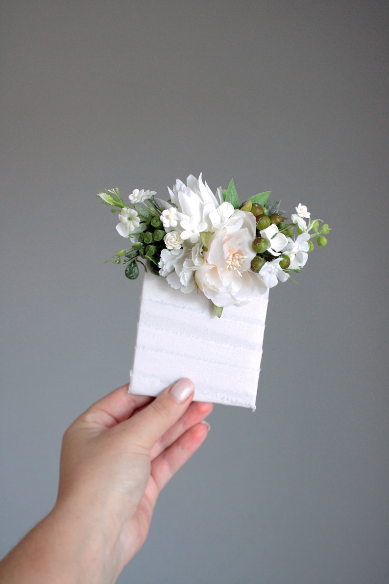 May include: A small bouquet of white flowers with green leaves and berries, arranged on a white card with a textured surface.