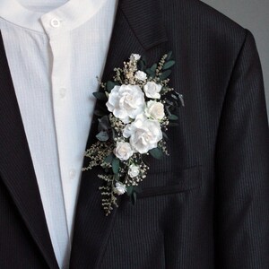 Close-up of white rose boutonniere with greenery and baby's breath on dark background.
