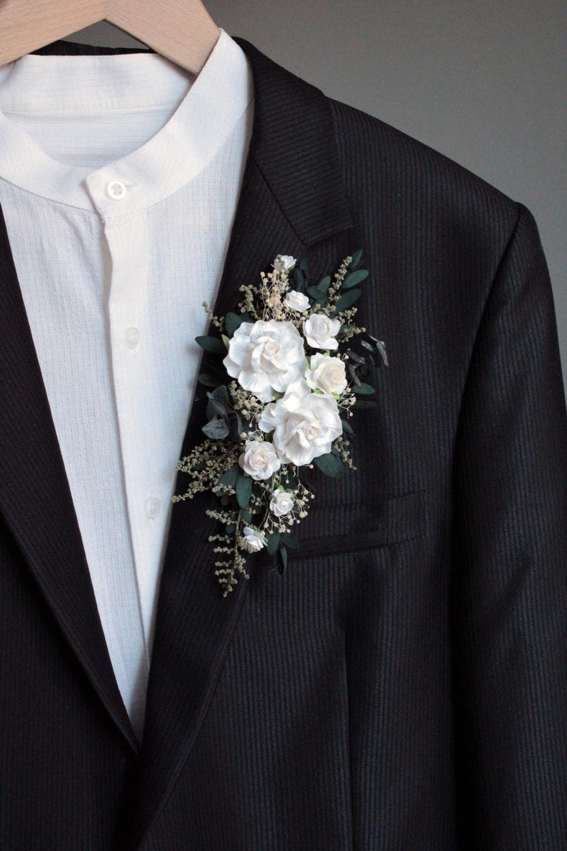 Close-up of white rose boutonniere with greenery and baby's breath on dark background.