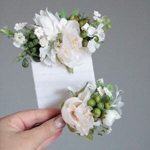 May include: Two white flower hair clips with green leaves and berries. The clips are on a white background.