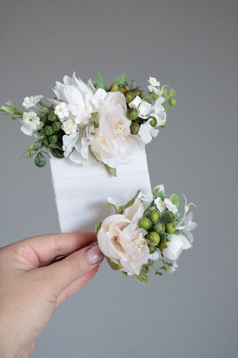May include: Two white flower hair clips with green leaves and berries. The clips are on a white background.