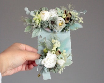 May include: A small bouquet of white flowers with green foliage and brown seed pods, arranged on a light blue fabric with a gold pin.