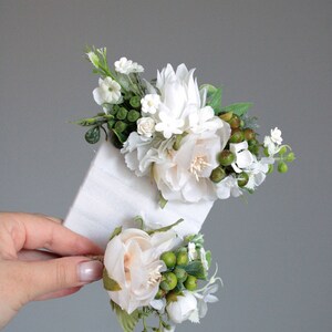 May include: Two white flower arrangements with green leaves and berries. The larger arrangement is on a white card and the smaller arrangement is held in a hand.