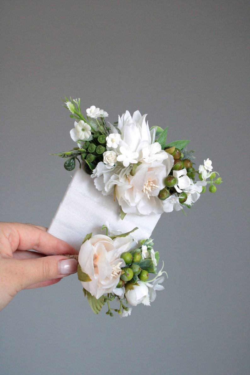 May include: Two white flower arrangements with green leaves and berries. The larger arrangement is on a white card and the smaller arrangement is held in a hand.