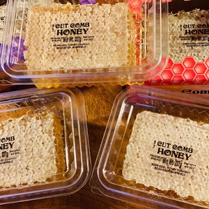 May include: Four clear plastic containers with lids, each containing a block of cut comb honey. The honey is a light golden colour and has a honeycomb texture. The containers are labelled "Cut Comb Honey" with the minimum weight of 227 grams. The honey is produced in England by Bear Cool Honey Company, Eckington, Sheffield.
