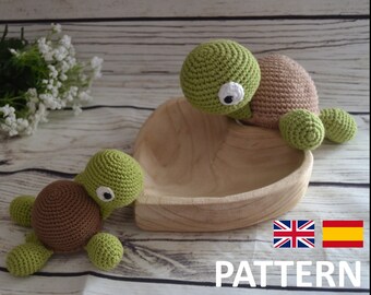 May include: Two crocheted turtle amigurumi toys, one brown and one green, sitting in a wooden heart-shaped bowl. The green turtle has a white eye and is sitting on the edge of the bowl. The brown turtle is sitting in the bottom of the bowl. The bowl is on a wooden surface.