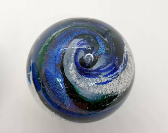 May include: A blue, green and white glass paperweight with a swirling pattern. The paperweight is round and has a shiny, iridescent finish.