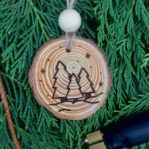 May include: A wood slice ornament with a wood-burned design of three pine trees and a moon. The ornament is hanging from a twine string with a wooden bead. The ornament is surrounded by green cedar branches.