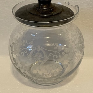 May include: A clear glass sugar jar with a silver lid and etched floral design. The lid has a small chip on the edge.