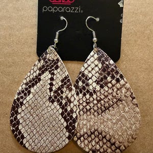 Może przedstawiać: Kolczyki w kształcie łezki ze wzorem skóry węża w odcieniach brązu i beżu. Kolczyki zawieszone są na srebrnych haczykach. Na czarnej etykiecie widnieje logo i nazwa marki "Paparazzi".