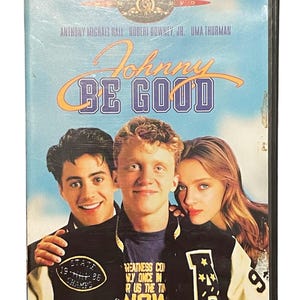 May include: DVD case for the film "Johnny Be Good" with Anthony Michael Hall, Robert Downey Jr., and Uma Thurman. The cover shows a blue sky background with the movie title in orange and white. The actors are wearing varsity jackets.