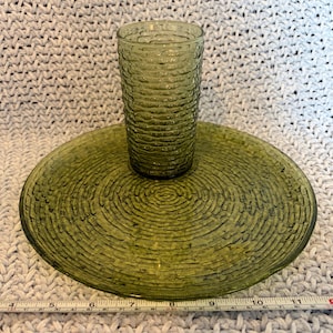 Puede incluir: Un juego de vaso y plato de vidrio verde. El vaso tiene un diseño texturizado y acanalado, y el plato tiene un patrón de círculos concéntricos. El conjunto es de color verde oliva claro y parece ser cristalería vintage.