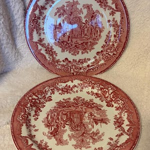 May include: Two antique ceramic plates with a red and white toile pattern. The plates feature a detailed landscape scene surrounded by floral borders. A ruler is placed near the plates for size reference.