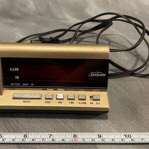 May include: A gold-colored digital clock with a black face and red display. The clock has a Sunbeam logo and buttons for snooze, hour, minute, time, alarm, on, and off. The clock is plugged in with a black cord.