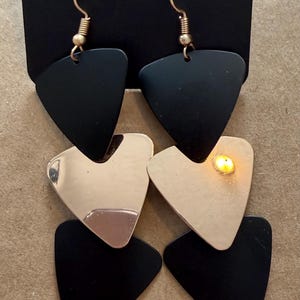 Könnte beinhalten: Schwarz-goldene geometrische Ohrringe. Die Ohrringe haben ein geschichtetes Design aus schwarzen und goldfarbenen Dreiecken. Sie hängen an einer schwarzen Karte mit dem Aufdruck "paparazzi".