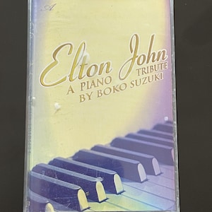 May include: A cassette tape case with a yellow background and a picture of a piano keyboard. The text on the case reads "Elton John A Piano Tribute By Boko Suzuki".