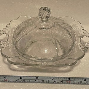 Puede incluir: Un plat de mantequilla de vidrio transparente con tapa y dos asas. El plato tiene un borde festoneado y un patrón floral.