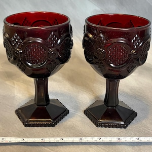 Pode incluir: Duas taças de vinho vintage vermelho rubi. As taças têm um padrão detalhado ao redor da taça e uma base hexagonal. A haste é de cor vermelho escuro. As taças têm aproximadamente 15 cm de altura.
