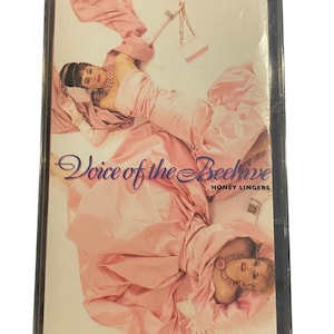 May include: A cassette tape with a pink and white design. The front of the tape features two women wearing pink dresses. The text "Voice of the Beehive" and "Honey Lingers" is printed on the tape.