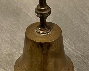 Pode incluir: Um pequeno sino de latão antigo com uma base arredondada e uma longa alça. O sino tem uma aparência envelhecida e manchada, sugerindo que é vintage. A alça é de um tom mais escuro de latão. O sino tem aproximadamente 10 cm de altura.