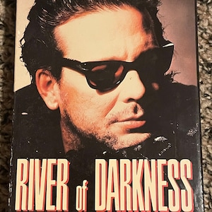 May include: A VHS video cassette cover featuring Mickey Rourke, Joe Pesci, and Theresa Russell in the film "River of Darkness". The cover features a close-up of Mickey Rourke wearing sunglasses and a dark jacket. The title "River of Darkness" is in large, red letters.