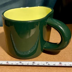 Puede incluir: Una taza de cerámica verde oscuro con interior amarillo brillante. La taza tiene un asa redondeada y una forma ligeramente irregular. La taza mide aproximadamente 7,6 cm de alto.